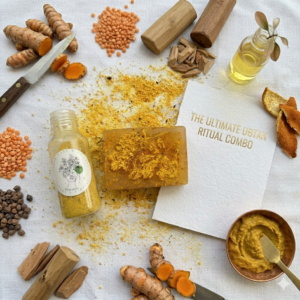 A luxury duo set featuring the 120g rectangular Brighten & Buff soap and a round, textured Ubtan scrubber. The products are displayed on a piece of raw burlap next to a pile of Channa Dal and dried rose petals, emphasizing the handcrafted, kitchen-to-skin nature of the kit.