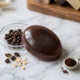 handcrafted, rectangular coffee soap bar with a rustic texture, featuring visible dark coffee grounds embedded throughout. The bar is photographed in warm, cinematic lighting on a wooden surface, surrounded by scattered whole roasted coffee beans and a hint of soft morning shadows.