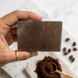 A handcrafted, rectangular coffee soap bar with a rustic texture, featuring visible dark coffee grounds embedded throughout. The bar is photographed in warm, cinematic lighting on a wooden surface, surrounded by scattered whole roasted coffee beans and a hint of soft morning shadows.