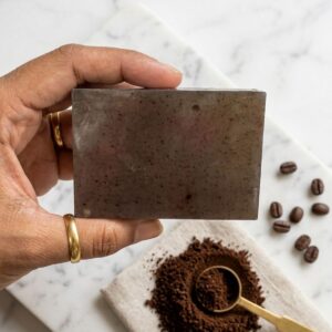 A handcrafted, rectangular coffee soap bar with a rustic texture, featuring visible dark coffee grounds embedded throughout. The bar is photographed in warm, cinematic lighting on a wooden surface, surrounded by scattered whole roasted coffee beans and a hint of soft morning shadows.