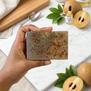 A close-up shot of a creamy, caramel-toned soap bar with soft, rounded edges, resting on a rustic stone soap dish. The bar has a slightly textured surface showing the natural fibers of fresh fruit pulp. In the background, a halved chiku fruit with its dark seeds and grainy brown flesh is softly blurred under warm, natural sunlight.