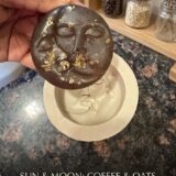 A beautifully handcrafted, crescent moon-shaped soap bar resting on a piece of dark, polished slate. The bar, named 'The Moon Bar,' has a deep caramel and espresso hue, and its textured surface is visibly speckled with rugged coffee grounds and rolled oats, resembling lunar craters and landscape.