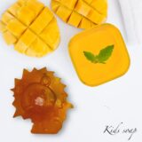Several small, cube-shaped mango soaps in a soft golden-orange color, arranged neatly in a minimalist ceramic bowl. The soaps have a smooth, matte finish with tiny visible flecks of real mango pulp.