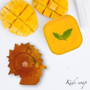 Several small, cube-shaped mango soaps in a soft golden-orange color, arranged neatly in a minimalist ceramic bowl. The soaps have a smooth, matte finish with tiny visible flecks of real mango pulp.