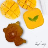 A vibrant and playful collection of small, mango-shaped soap bars in bright golden-orange hues. The soaps are arranged whimsically on a white tiled surface with scattered soap bubbles and a few fresh mango cubes nearby. The lighting is bright and cheerful, highlighting the smooth, "pop-style" shapes that are perfectly sized for small hands.
