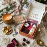An open Sweet Sisterhood Rakhi gift box neatly displaying a round Petal and Grain rose moon soap, a small jar of Manjistha lip balm, an organic ubtan scrubber, and aesthetic dried floral accents.
