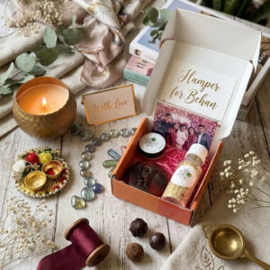 An open Sweet Sisterhood Rakhi gift box neatly displaying a round Petal and Grain rose moon soap, a small jar of Manjistha lip balm, an organic ubtan scrubber, and aesthetic dried floral accents.