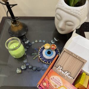 Centered is a beaded evil eye coaster with two small traditional katoris (bowls) holding roli and chawal. To the right is an open orange gift box containing purple shred, a 'Happy Raksha Bandhan' greeting card with a red and yellow thread rakhi, a small gold bar, and a wooden photo frame etched with 'YOU'RE THE BEST' and 'Brother EVER'.